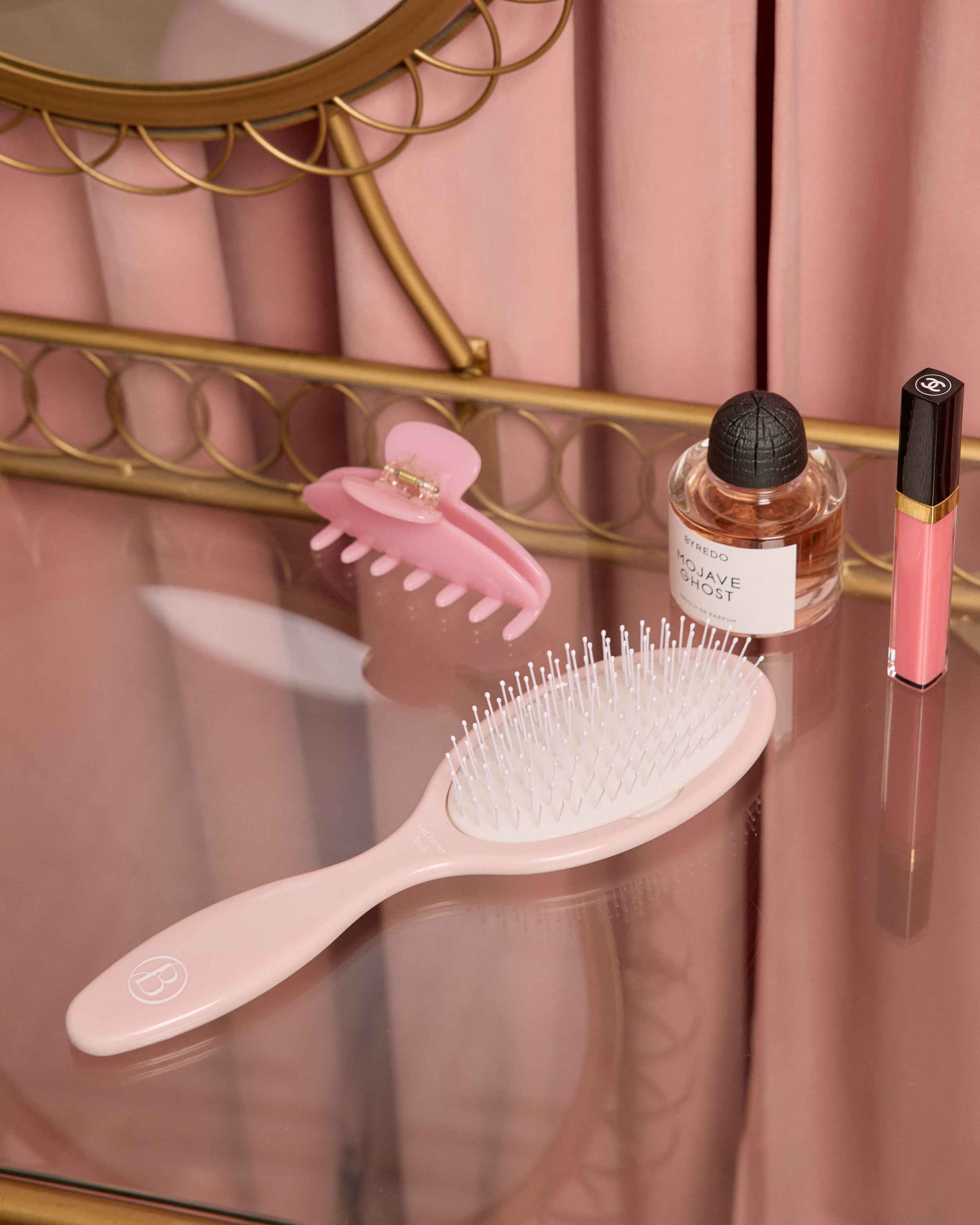 Pink hairbrush, hair clip, and lip gloss on a glass vanity table with a decorative mirror in the background.