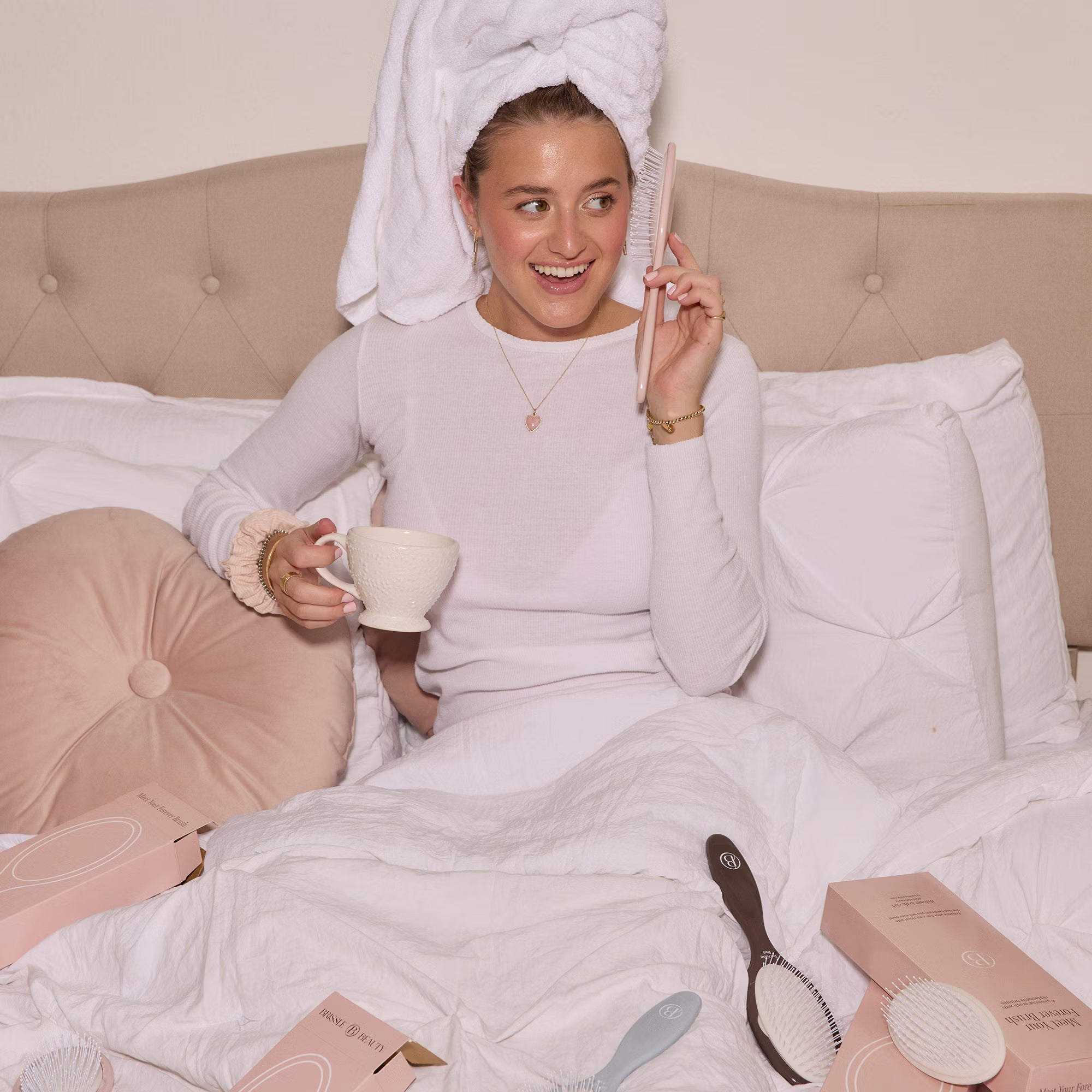 Woman in a white clothes with a towel on her head, holding a mug, sitting on a bed with hair care products around and holding a hairbrush as a telephone.